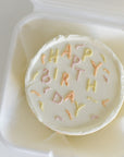 Round white cake with 'Happy Birthday' text in colorful icing, placed in a clear container.