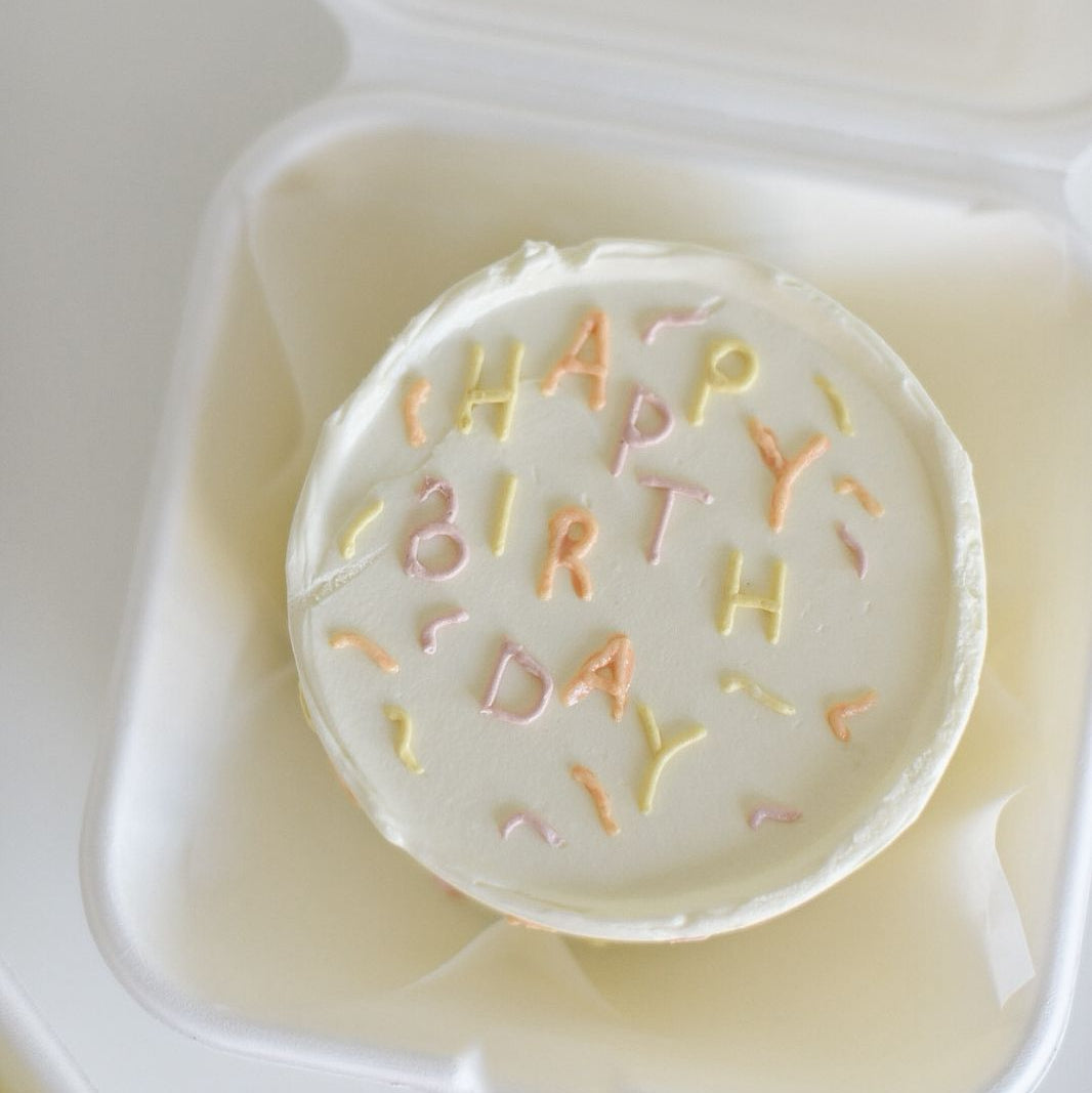 Round white cake with 'Happy Birthday' text in colorful icing, placed in a clear container.