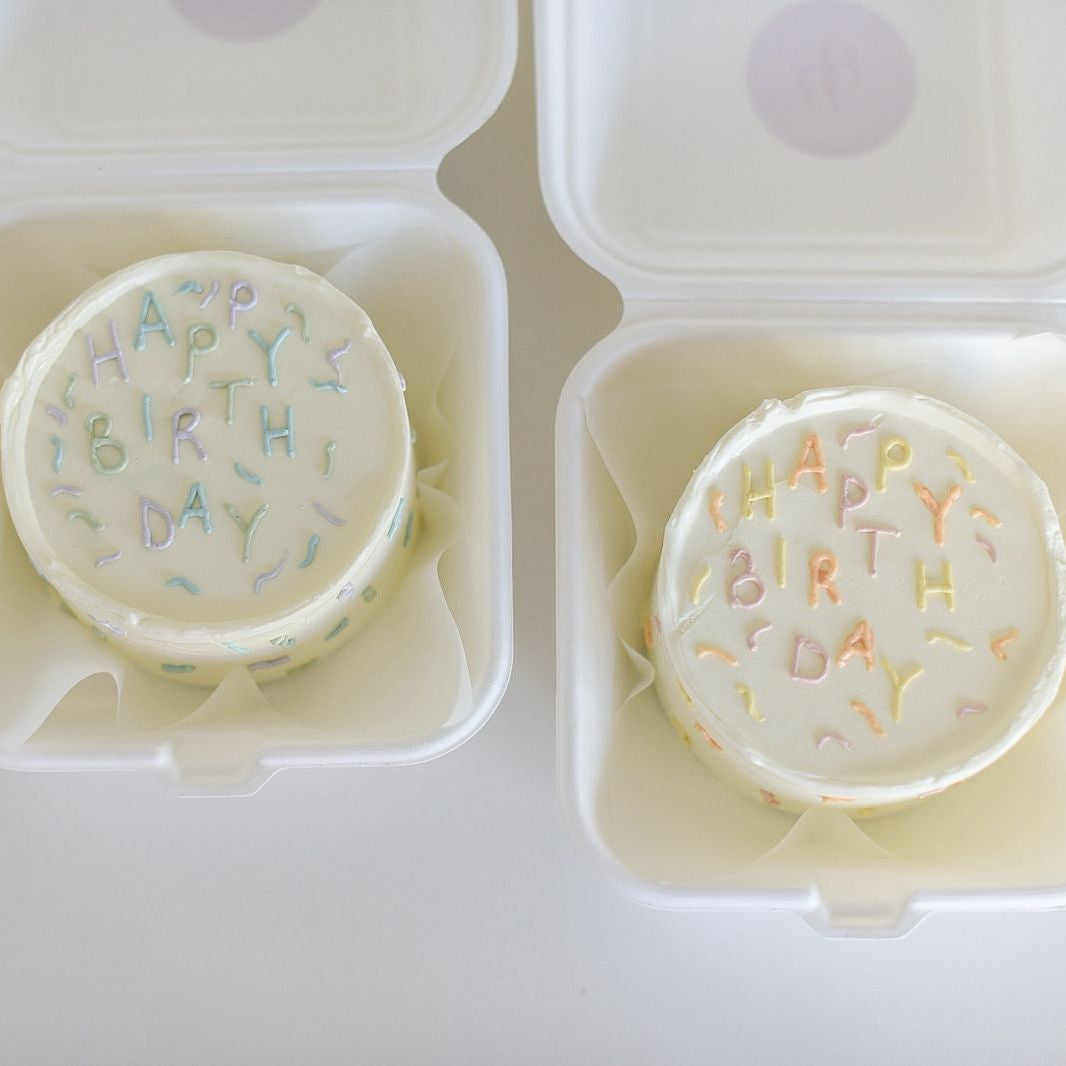 Two round cookies with 'Happy Birthday' sprinkles in white containers on a light background