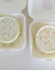 Two round cookies with 'Happy Birthday' sprinkles in white containers on a light background